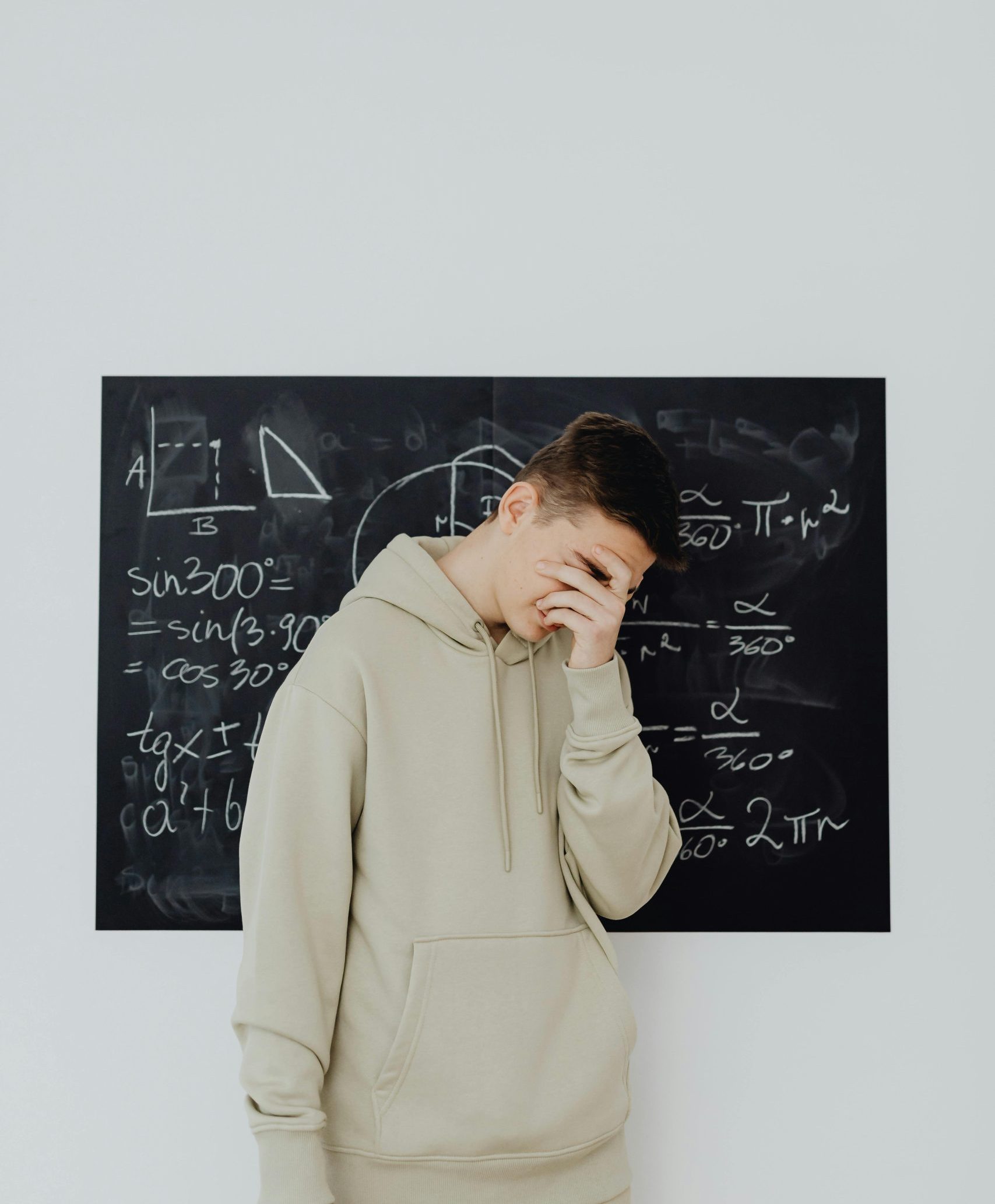 A young boy in a hoodie stands covering his face in frustration before a math equation-filled chalkboard.